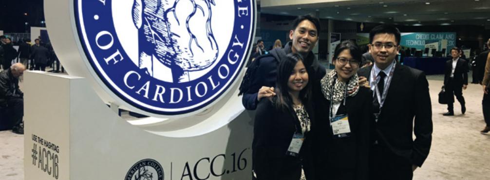 Four people stand smiling in front of an illuminated American College of Cardiology sign in a conference hall.